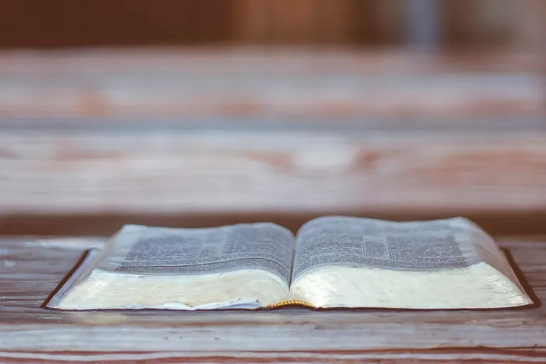 An open Bible rests on a wooden church pew, with the empty interior softly out of focus.